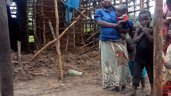 Frau mit Baby auf dem Arm steht vor zerstörter Hütte.