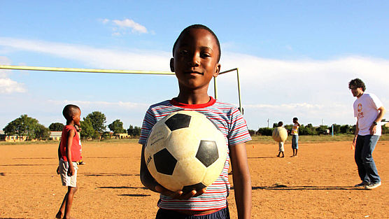 Ein Junge hält einen Fußball in der Hand, im Hintergrund wird gekickt