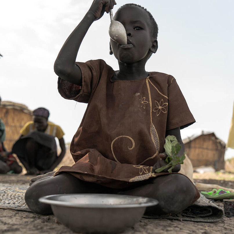 Die fünfjährige Nyahok sitzt auf dem Boden und isst aus einem tiefen Teller, der vor ihr steht.