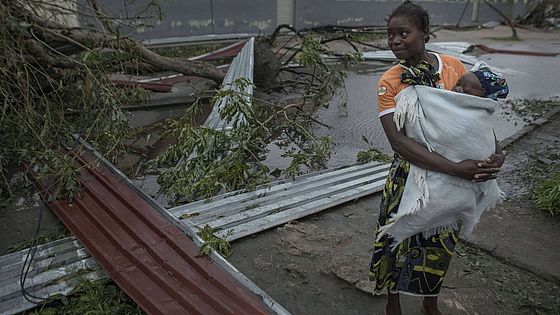 Frau mit Baby in einer Trage steht vor einer zerstörten Schule.