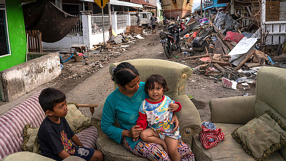 Eine Frau sitzt mit zwei Kindern auf Sofas auf der Straße, nachdem ihr Haus vom Tsunami zerstört wurde. Im Hintergrund ist ein gestrandetes Schiff zu sehen.