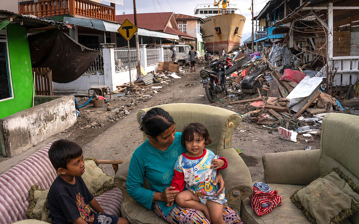 Eine Frau sitzt mit zwei Kindern auf Sofas auf der Straße, nachdem ihr Haus vom Tsunami zerstört wurde. Im Hintergrund ist ein gestrandetes Schiff zu sehen.