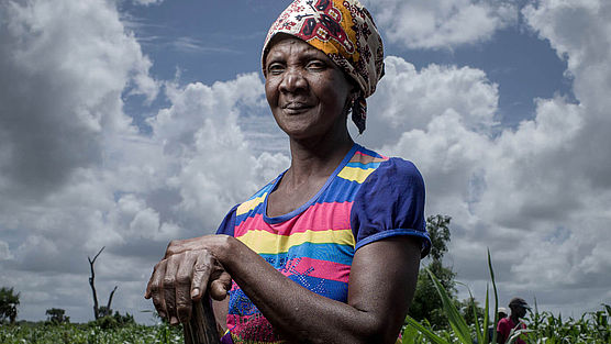 Die70-jährige Mantando Joan steht in bunter Kleidung mit einem Spaten in der Hand auf einem Feld