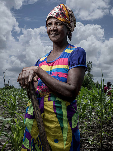 Die70-jährige Mantando Joan steht in bunter Kleidung mit einem Spaten in der Hand auf einem Feld
