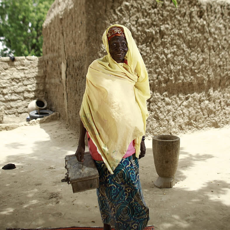 Schatzmeisterin Zeinabou steht mit der Geldkiste in der Hand vor einem Steinhaus.