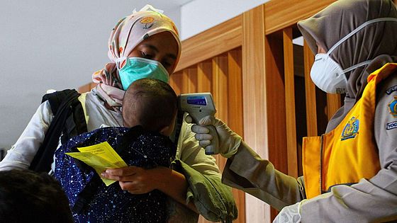 Eine Frau mit Maske misst die Temperatur eines Babys, das von seiner Mutter in einer Trage getragen wird.