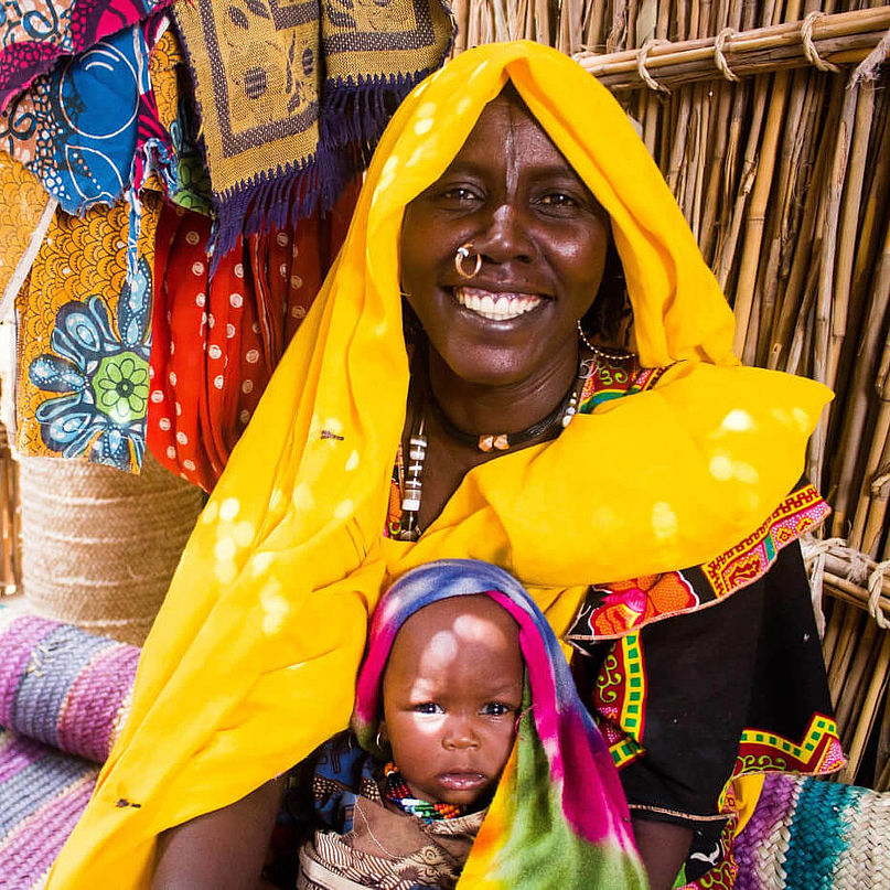 In einem Flüchtlingscamp im Tschad hat eine junge Mutter in gelber Kleidung lächelnd ihr Baby auf dem Schoß.