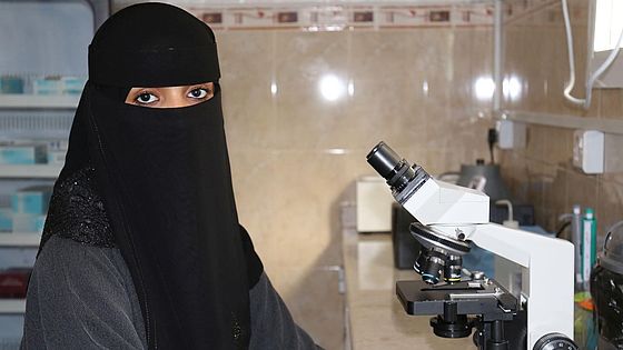 Heba aus dem Jemen schaut in die Kamera, vor ihr steht ein Mikroskop.