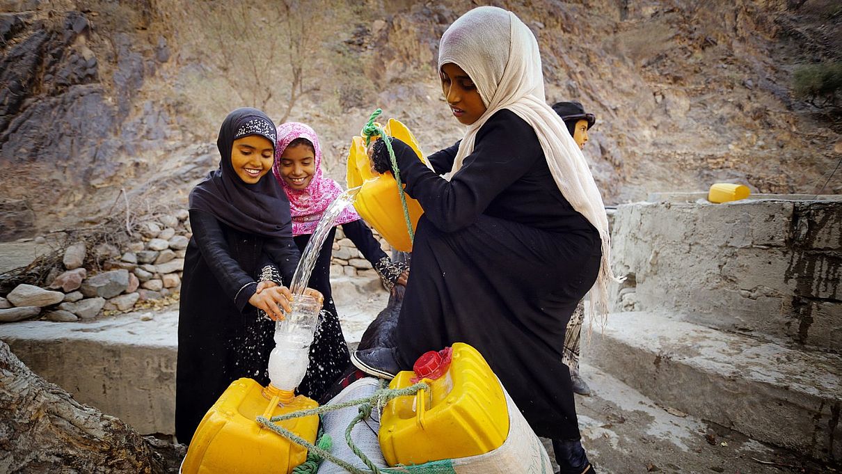 Drei Mädchen zapfen von einem Brunnen Wasser ab.
