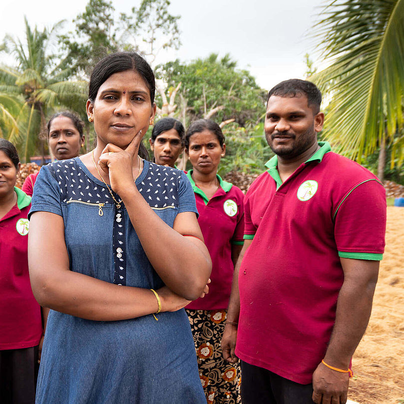 Unternehmerin Sharmini trägt ein blaues Kleid, im Hintergrund stehen ihre Mitarbeitenden