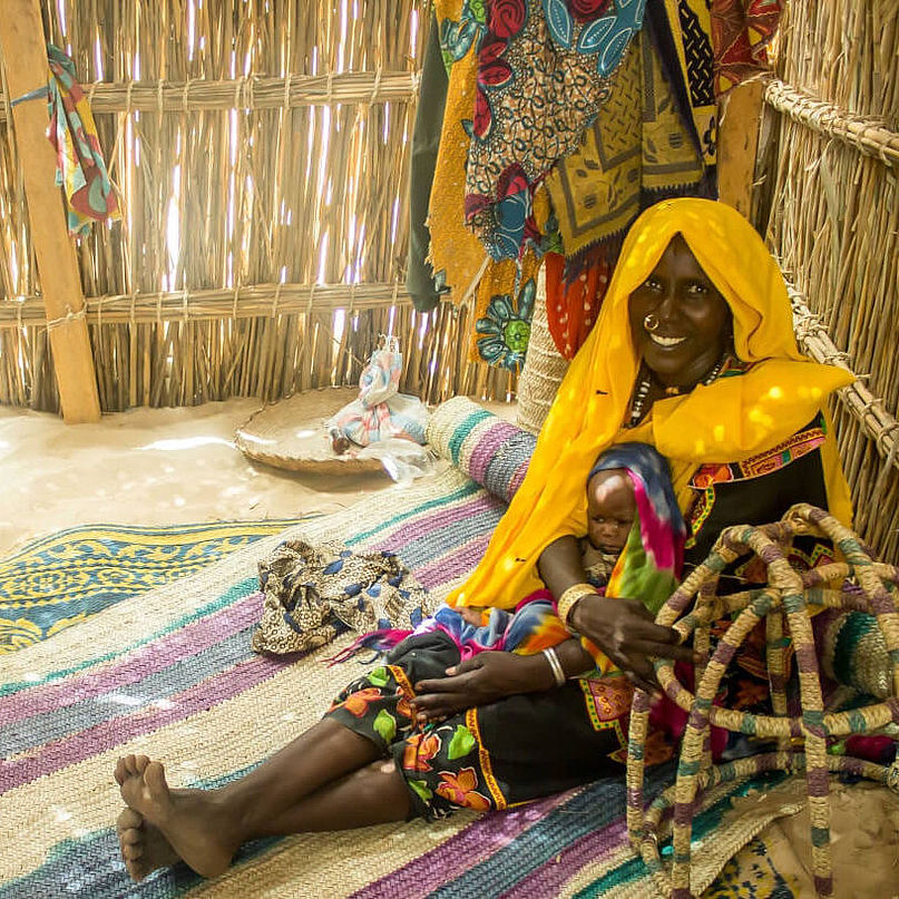 Eine Frau in bunter Kleidung sitzt lächelnd mit ihrem Baby auf dem Schoß in ihrem neuen Zuhause in einem Flüchtlingscamp im Tschad