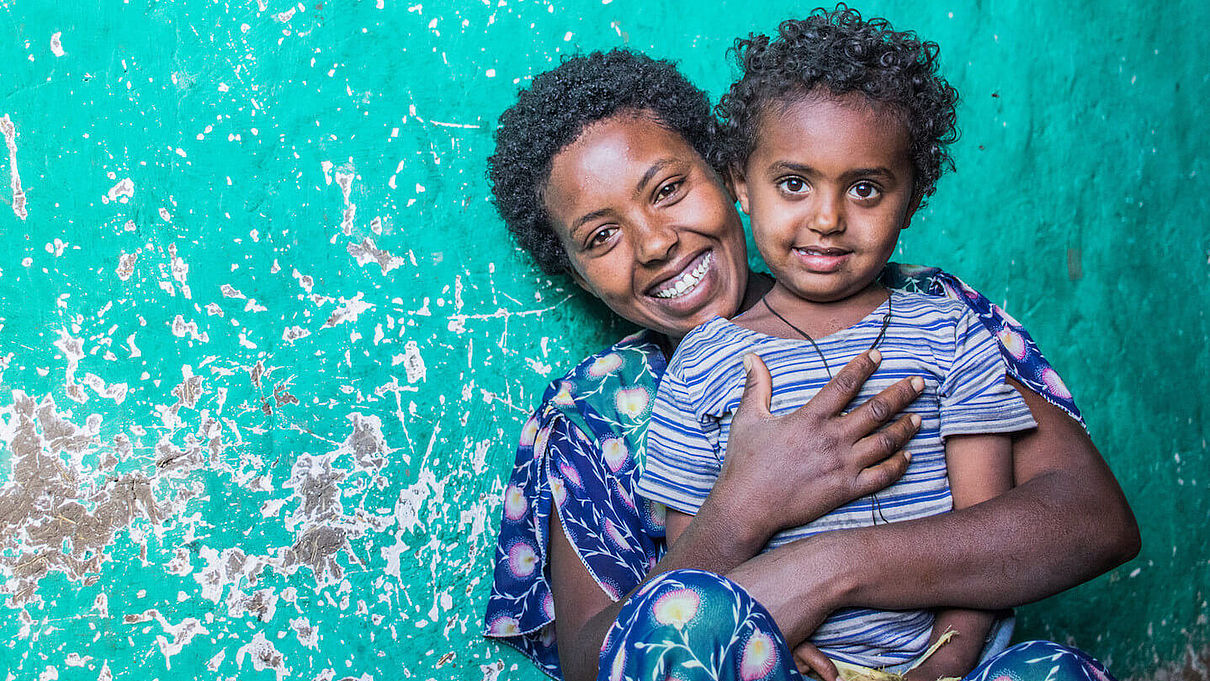 Mutter und Tochter aus Äthiopien sitzen lächelnd vor einer türkisfarbenen Wand.