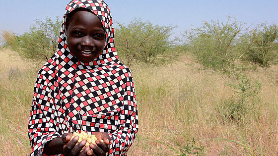 Die zehnjährige Zahara aus dem Niger steht in buntgemusteter Kleidung auf einem Feld, hält Erdnüsse in den Händen und lächelt.