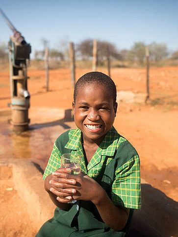 Ein Mädchen sitzt vor einem Wasserbrunnen und hält ein Glas mit sauberem Trinkwasser in den Händen