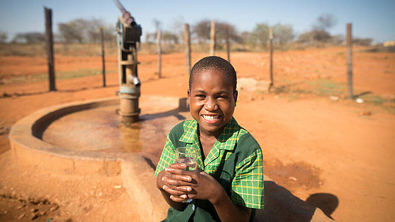 Ein Mädchen sitzt vor einem Wasserbrunnen und hält ein Glas mit sauberem Trinkwasser in den Händen