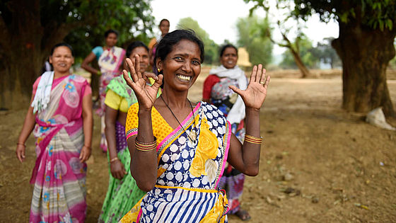 Dilmani Kujur in einem bunt gemusterten Kleid formt mit ihren Händen das 1,5-Grad-Zeichen, im Hintergrund stehen weitere Frauen.