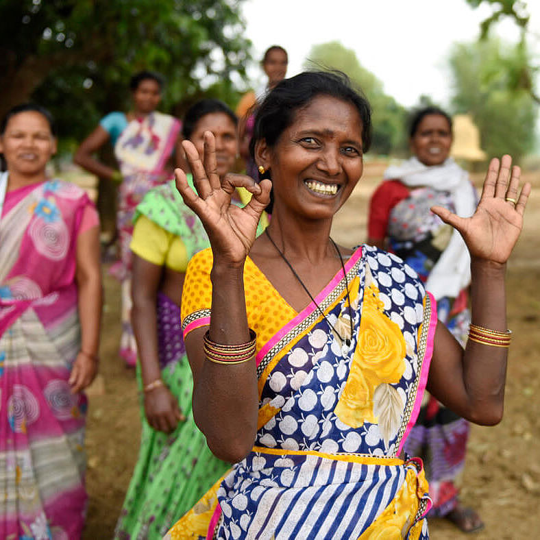 Dilmani Kujur in einem bunt gemusterten Kleid formt mit ihren Händen das 1,5-Grad-Zeichen, im Hintergrund stehen weitere Frauen.