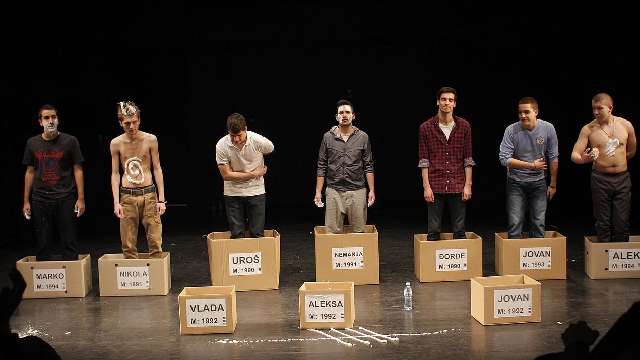 Aufführung eines Theaterstücks Mehrere Männer stehen in Kartons auf einer Bühne.
