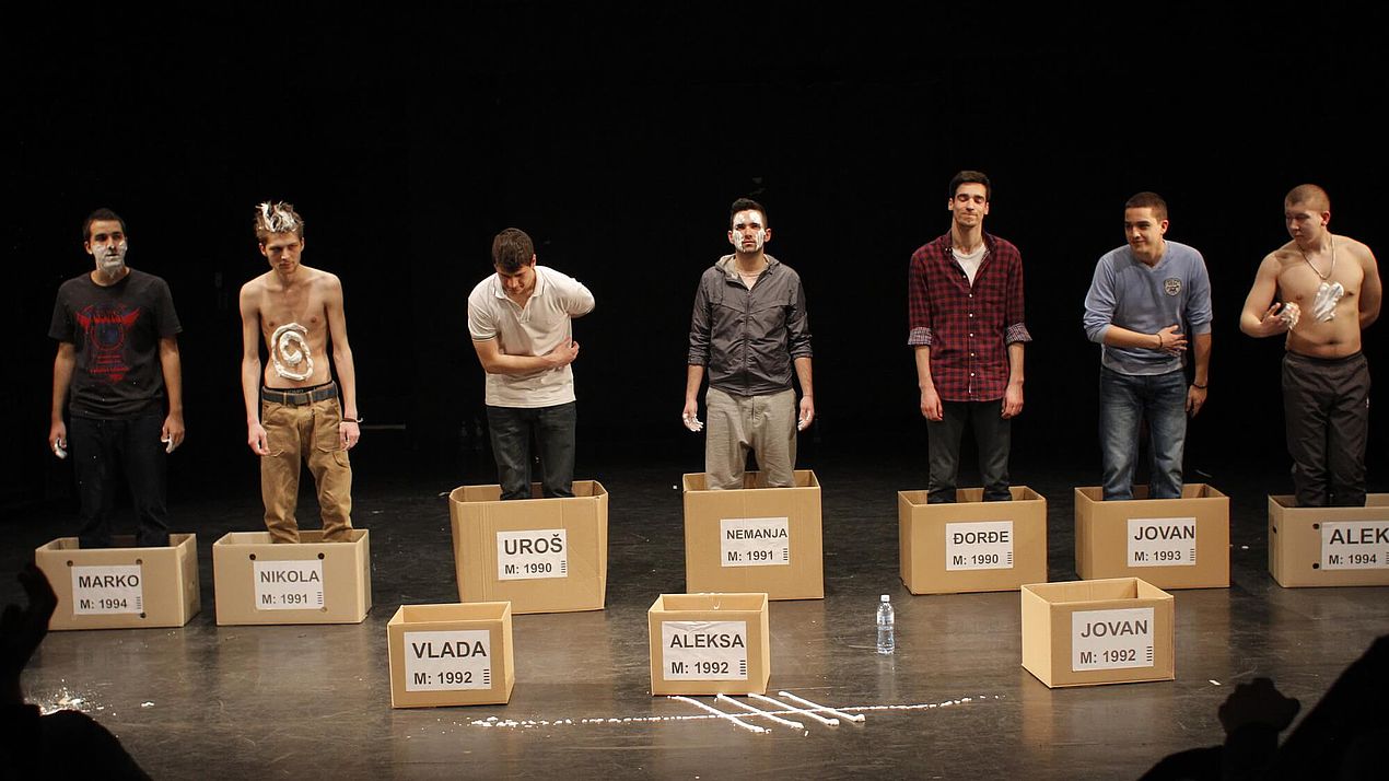 Aufführung eines Theaterstücks Mehrere Männer stehen in Kartons auf einer Bühne.