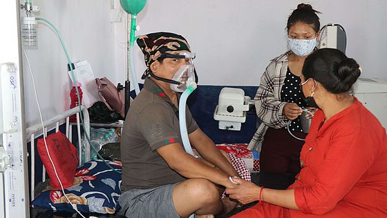 Ein Mann mit einer Sauerstoffmaske im Krankenhaus in Nepal, an seinem Bett sitzen zwei Frauen