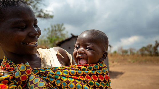 Eine Mutter trägt ihr Baby in einem bunten Tuch, beide lachen