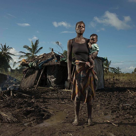 Frau steht mit Baby auf dem Arm vor zerstörter Hütte in Mosambik..