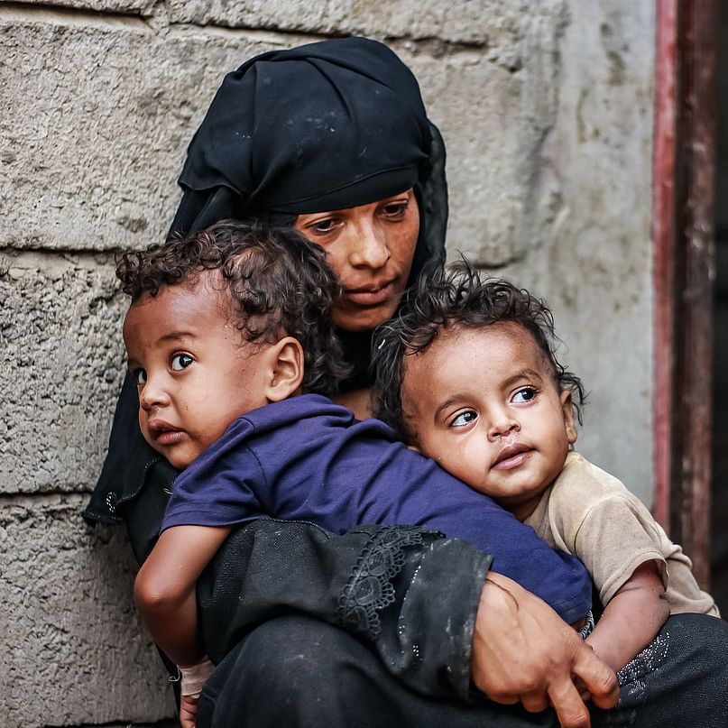 Eine Frau sitzt mit zwei Kindern auf dem Schoß vor einer Wand und sieht traurig aus.