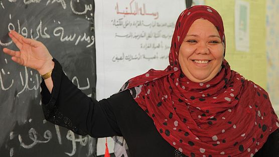 Eine Lehrerin mit rotem Kopftuch zeigt lächelnd auf eine Tafel.