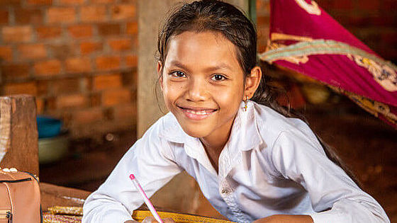 Ein Mädchen sitzt mit einem Stift in der Hand an einer Schulbank und lächelt