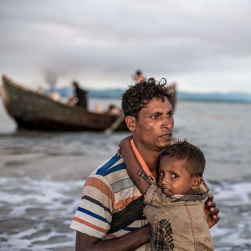 Ein geflüchteter Vater aus Myanmar trägt sein Kind in Bangladesch ans Ufer.