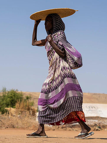 Eine Frau geht und trägt eine runde Strohmatte über dem Kopf, im Hintergrund sieht man Zelte des Flüchtlingscamps