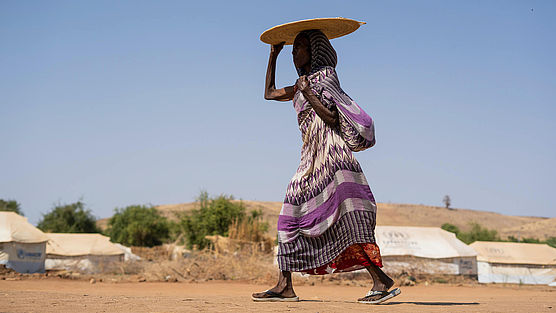 Eine Frau geht durch ein Flüchtlingscamp und hält sich etwas über den Kopf, um Schatten zu spenden