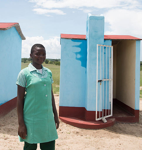 Ein Mädchen in Schuluniform in Simbabwe, im Hintergrund sieht man zwei mädchenfreundliche Toiletten