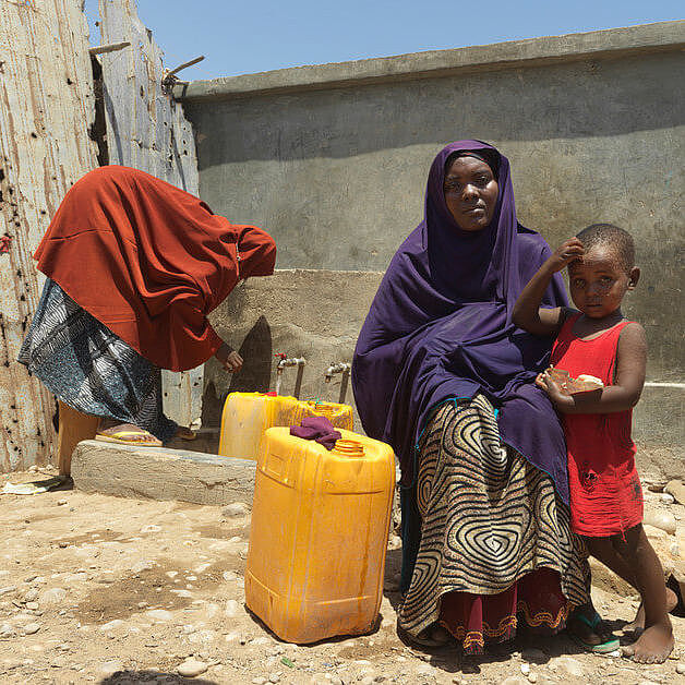 Layla sitzt mit ihrem Kind vor der Wasserstelle in Bosaso, Somalia, und hält einen gelben kanister.