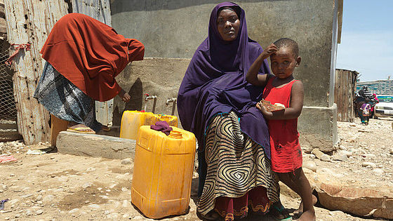 Layla sitzt mit ihrem Kind vor der Wasserstelle in Bosaso, Somalia, und hält einen gelben kanister.