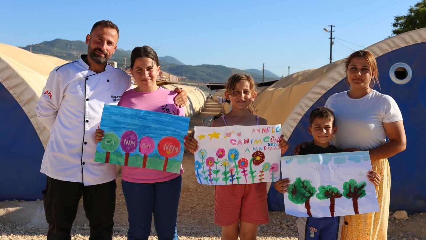 Drei Kinder stehen mit ihren Eltern vor ihrer Notunterkunft und halten selbst gemalte Bilder in die Kamera.